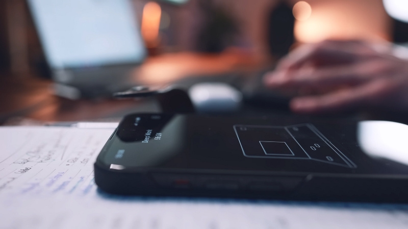 A Smartphone with A Timer App Open Resting on A Notebook, with A Person Working on A Laptop in The Background